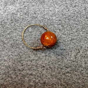 Puede incluir: Un anillo hecho a mano con una banda dorada y una gran cuenta redonda de color ámbar. La cuenta tiene una superficie texturizada y está sujeta a la banda con alambre. El anillo está sobre un fondo gris.