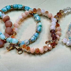 May include: Four beaded bracelets in various colors. One bracelet features large, round, pinkish-brown beads. Another has turquoise and brown beads. The third has peach and clear beads. The last has clear and light blue beads.