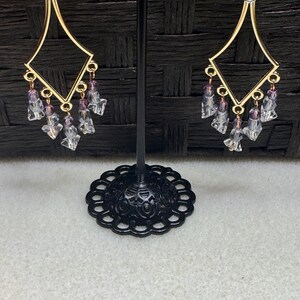 Könnte beinhalten: Goldfarbene Chandelier-Ohrringe mit geometrischem Design. Jeder Ohrring hat einen rautenförmigen Rahmen mit herabhängenden Clustern aus klaren und lila facettierten Perlen. Die Ohrringe werden auf einem schwarzen Ständer vor dunklem Hintergrund präsentiert.