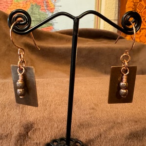 May include: A pair of copper earrings with a rectangular, dark brown pendant. The earrings feature a coiled wire and two decorative beads. The ear hooks are copper-colored. The earrings are displayed on a black stand against a brown background.