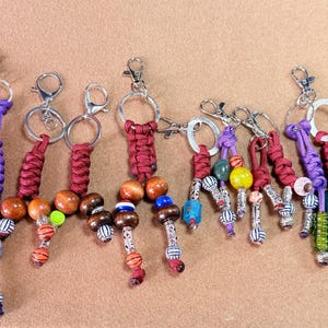 May include: A collection of ten handmade keychains featuring braided paracord in purple and maroon, accented with various wooden, plastic, and metal beads. Each keychain includes a silver or gold-toned metal lobster clasp and a circular ring.