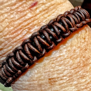 May include: A dark brown braided bracelet. The bracelet is made of tightly woven, round cords, creating a textured surface. The bracelet is a rich, warm brown color, and appears to be made of leather or a similar material. The design is simple yet stylish.