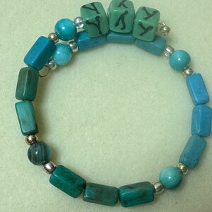 May include: A turquoise and teal beaded bracelet. The bracelet features a mix of round and rectangular beads, with silver-colored spacers. Three green square beads with black letter "Y" are at the top. The bracelet is on a light green surface.