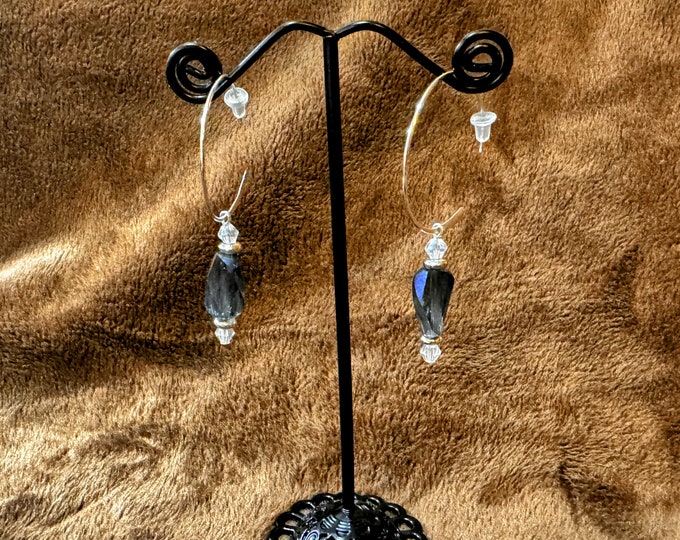 Handmade 2 1/4” round silver sideways hoop earrings with a twisted crystal bead in blue hues hanging from it.