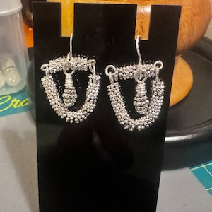 May include: A pair of silver-toned earrings displayed on a black stand. Each earring features a beaded, teardrop-shaped design with a central, spiraled element. The earrings are intricate and detailed, showcasing a handcrafted aesthetic.