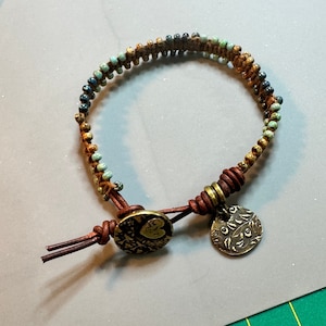 Könnte beinhalten: Handgefertigtes Armband mit einem gewebten Muster aus kleinen, farbigen Perlen in Türkis, Braun und Gold. Es hat einen Lederverschluss mit einem dekorativen Metallanhänger und einen runden Knopfverschluss. Ein einzigartiger Schmuck.