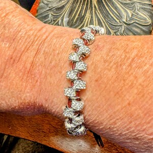 May include: A silver bracelet with clear, sparkling stones and a red leather band. The bracelet features multiple silver rings, each adorned with small, glittering stones. The red band weaves through the rings, creating a stylish accessory.