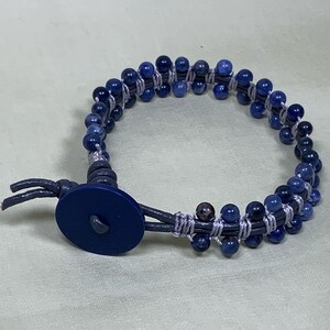 Peut inclure: Un bracelet enroulé avec des perles bleu foncé et une fermeture à bouton bleu marine. Les perles sont enfilées avec un fil gris clair, et le bracelet a une lanière en cuir. Le bracelet est conçu pour s'enrouler autour du poignet.