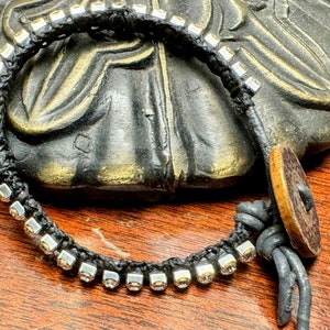 May include: Black braided bracelet with silver-colored square beads. The bracelet has a wooden button closure and is displayed on a dark surface. The bracelet is a handmade jewelry item.