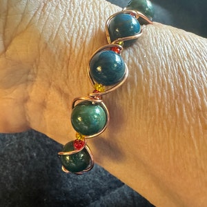 May include: A handmade bracelet featuring dark green and blue gemstone beads. The beads are wrapped in copper wire, with small red and yellow accent beads. The bracelet is shown on a wrist.