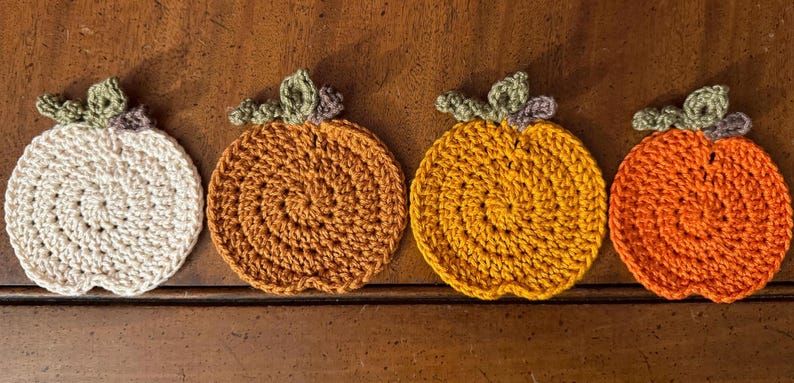 May include: Four crocheted pumpkin coasters in white, orange, brown, and yellow. The coasters are round and have a stem with leaves on top.