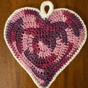 May include: A crocheted heart-shaped dishcloth in shades of pink and purple with a white border and a loop at the top.
