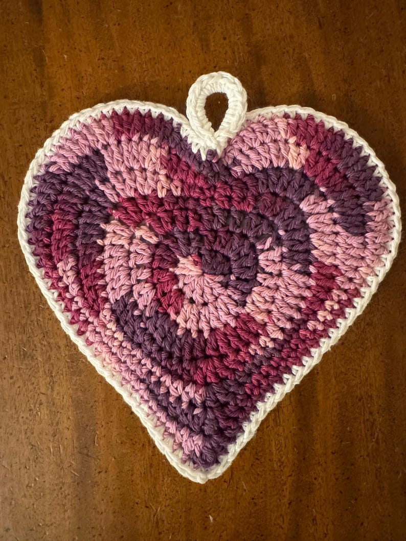 May include: A crocheted heart-shaped dishcloth in shades of pink and purple with a white border and a loop at the top.