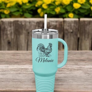Può includere: Un thermos da viaggio in acciaio inossidabile turchese con un design di gallo nero e il nome "Melanie" stampato sul lato. Il thermos ha un manico e una cannuccia.