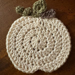 May include: A crocheted pumpkin coaster in cream with green and brown leaves.