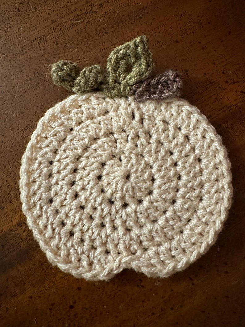 May include: A crocheted pumpkin coaster in cream with green and brown leaves.