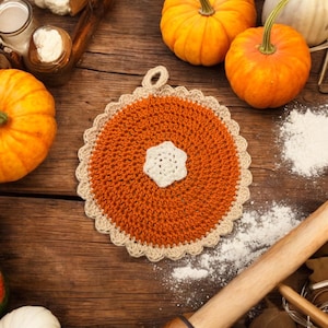 May include: A crocheted pumpkin pie dish trivet with a white center and orange crocheted edges. The trivet is on a wooden surface with pumpkins and flour.