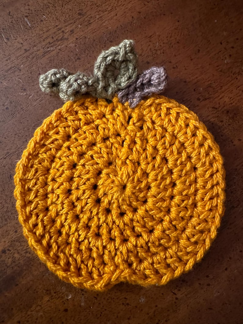 May include: A crocheted pumpkin coaster in orange with green and purple leaves.