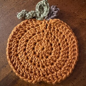 May include: A crocheted pumpkin coaster in orange with green and brown leaves on top.