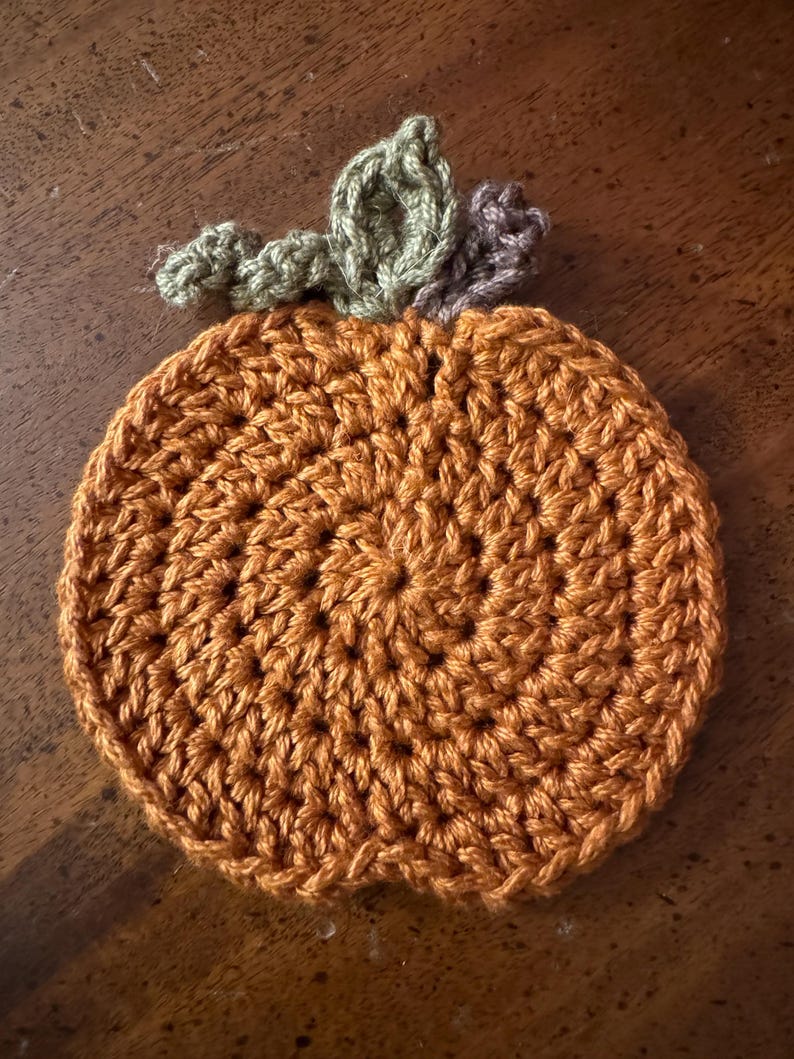 May include: A crocheted pumpkin coaster in orange with green and brown leaves on top.