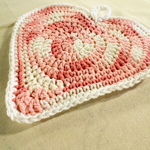 May include: A pink and white crocheted heart-shaped pot holder with a loop for hanging.