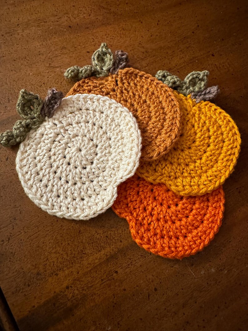 May include: Four crocheted pumpkin coasters in shades of orange, yellow, and white. The coasters are arranged in a stack, with the white coaster on top.