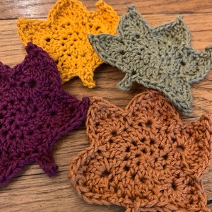 May include: Four crocheted leaf-shaped coasters in different colors: mustard yellow, olive green, burgundy, and burnt orange.