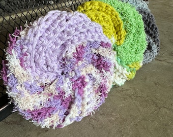 May include: A set of four circular, hand-crocheted scrubbers arranged in a row. The first is a variegated blend of lavender, purple, and cream. The second is a solid chartreuse, the third is a textured light green, and the fourth is dark gray.