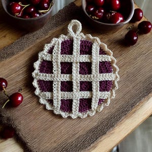 May include: A crocheted pot holder shaped like a cherry pie with a brown crust and a burgundy filling. The pot holder has a loop handle.