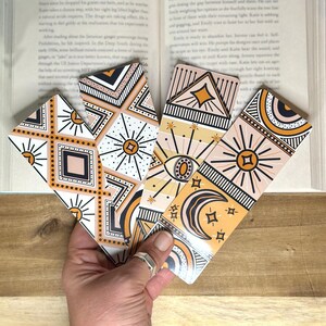 May include: Four colorful bookmarks with geometric patterns and celestial designs. The bookmarks feature a mix of orange, yellow, black, and white colors. The designs include stars, moons, eyes, and geometric shapes.