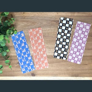 May include: Four different patterned bookmarks. One bookmark has a blue and white checkered pattern with dripping blue paint. Another bookmark has a pink and white checkered pattern with dripping orange paint. A third bookmark has a black and white checkered pattern with gold stars. The final bookmark has a pink and white checkered pattern with white hearts.
