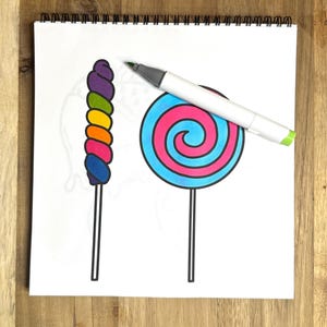 May include: A white sheet of paper with a spiral binding at the top. A rainbow swirl lollipop and a blue and pink swirl lollipop are drawn on the paper. A gray marker is lying on the paper.
