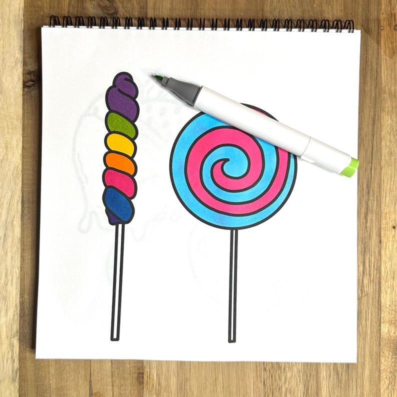 May include: A white sheet of paper with a spiral binding at the top. A rainbow swirl lollipop and a blue and pink swirl lollipop are drawn on the paper. A gray marker is lying on the paper.