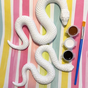 May include: Two white, textured snake figurines with detailed scales. The set includes three small circular containers of paint in brown, white, and black, along with a single paintbrush featuring a blue handle and a yellow-tipped bristle head.