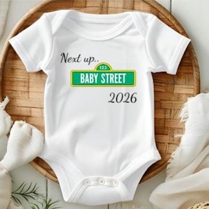May include: White baby onesie with the text "Next up.. BABY STREET 2026" in black and green. The onesie is on a woven basket with a stuffed bunny and a white cloth.