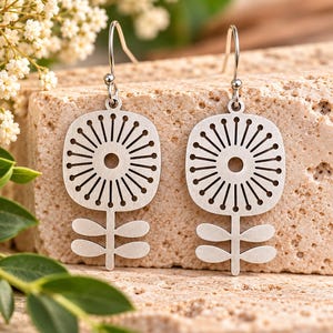 May include: A pair of silver-toned metal drop earrings featuring a flat, circular flower design with radiating lines and a central cutout. Each earring has a thin stem with two pairs of rounded leaves and a simple curved hook for attachment.