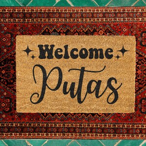 May include: A brown doormat with the text "Welcome Puta's" in black lettering. The doormat is surrounded by a red, black, and blue patterned border.