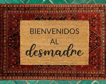 May include: A brown coir doormat with the text "BIENVENIDOS AL DESMADRE" in black lettering. The doormat is framed by a red, black, and white patterned rug.