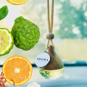 May include: A hanging car air freshener with a teardrop-shaped wooden cap and a clear glass base containing liquid. A round silver tag attached to the cap features the text AROMA. The background displays floating images of a lemon slice, bergamot, orange slice, tea bag, and wood shavings.