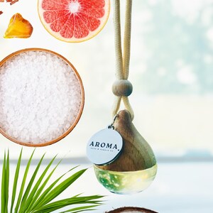 May include: A hanging glass car air freshener with a wooden cap and a round silver tag labeled AROMA. The bottle contains a clear liquid and is suspended by a beige cord. Surrounding elements include a grapefruit slice, sea salt, and coconut.