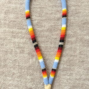 May include: A beaded lanyard with a light blue background and colorful stripes of red, orange, yellow, black, and white. The lanyard has a silver key ring attached at the bottom. The lanyard is made of small, round beads.