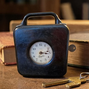 May include: A black vintage Simplex time clock with a handle, a round clock face, and a key. The clock face has black numbers and hands. A set of keys rests on the wooden surface in front of the clock.