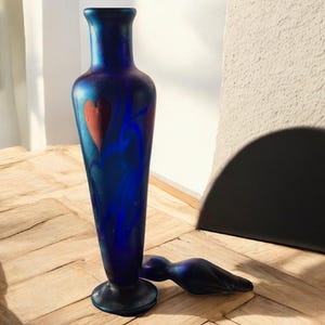 Può includere: Un vaso alto di vetro blu con base svasata e collo stretto. Il vaso presenta un motivo a cuore rosso e accenti blu a spirale. Un tappo di vetro abbinato è appoggiato su un lato. Il vaso è alto circa 25 cm.