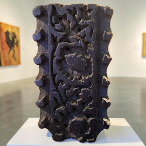 May include: A dark brown, rectangular wooden block with intricate floral carvings. The block has a textured surface and is displayed on a white pedestal in a gallery setting. The edges feature rounded protrusions.
