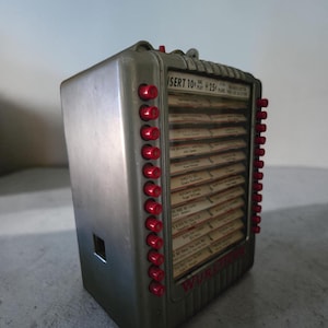 Könnte beinhalten: Vintage silberfarbene Jukebox mit roten Auswahltasten. Die Jukebox hat eine Anzeige mit Songtiteln und dem Text "INSERT 10¢ PLAY 25¢". Ein klassisches Stück Musikgeschichte.