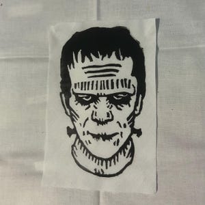 May include: A black and white linocut print of Frankenstein's monster on a white fabric square. The monster's face is detailed with a furrowed brow, stitched neck, and a stern expression. The artwork is centered.