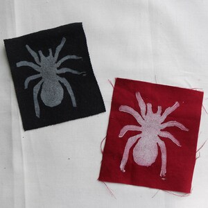 May include: Two fabric patches with a white spider silhouette. One patch is black and the other is red.