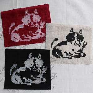 May include: Three fabric patches with a white devil cat design. One patch is red, one is black, and one is off-white. The devil cat has horns, a tail, and a pitchfork. The patches are on a white background.