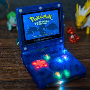 May include: A blue Game Boy Advance SP handheld gaming console with a bright, illuminated screen displaying the Pokemon Sapphire Version game. The console has a translucent blue case with a clear screen and buttons.