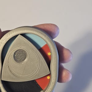 May include: A gray, 3D-printed, circular object with a triangular center. The triangle is divided into sections of black, red, and blue. Two yellow dots are visible. The object has a textured surface.